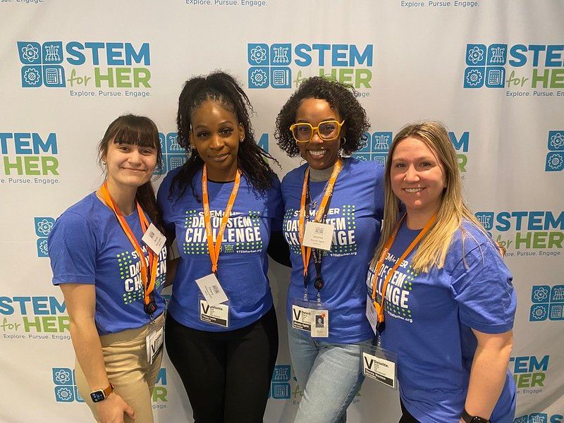 Four women in blue STEM shirts posing at a STEM-themed event backdrop.