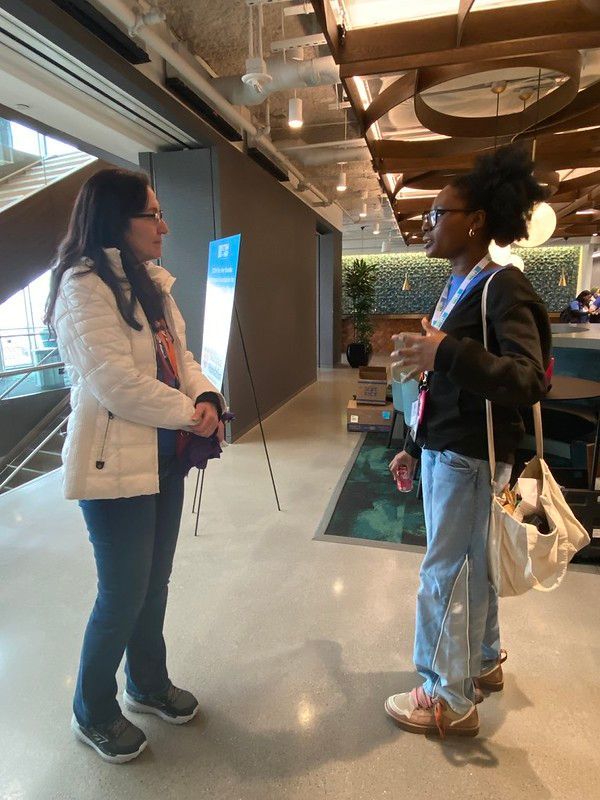 Two women talking in a modern indoor corridor, one in a white coat and one in a black jacket.