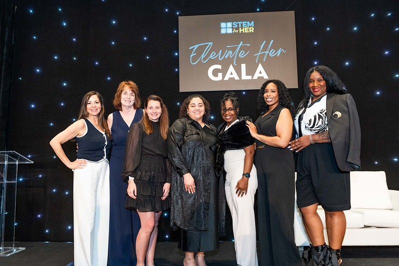 Six people pose onstage at a gala, smiling in black formal outfits beside a “Gala” sign.