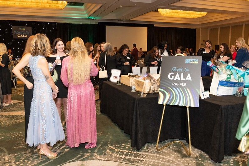 Guests mingle at a gala reception beside a table and sign under warm chandelier lighting.