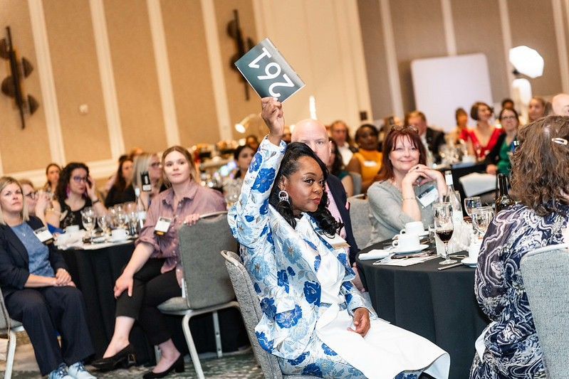 Audience seated at banquet tables in a conference hall, one woman raising a bid card.