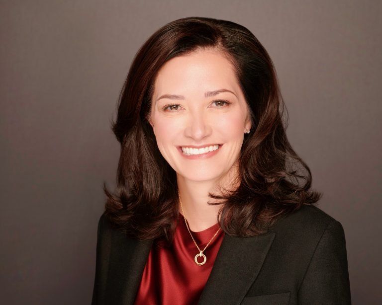 Smiling woman in a black blazer and red blouse against a plain background.