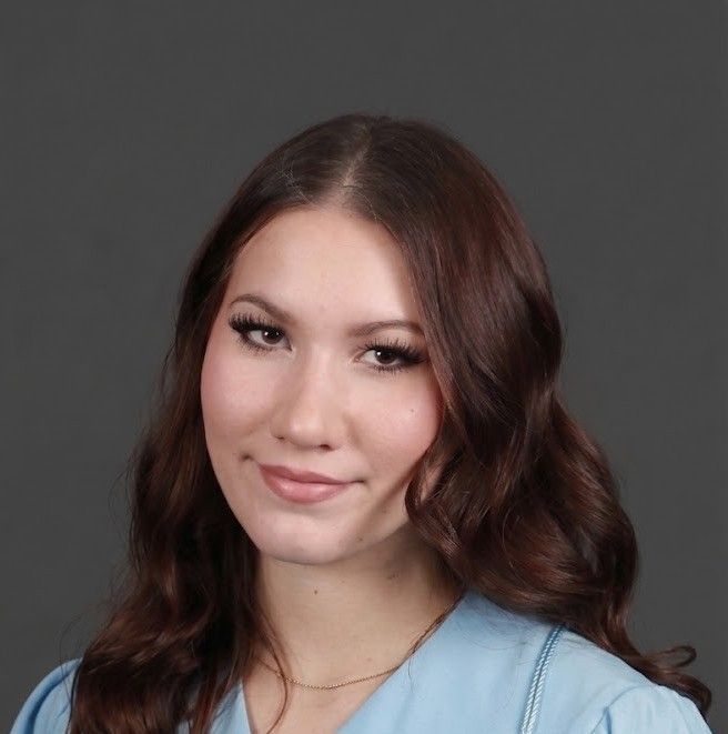 Portrait of a smiling woman with wavy brown hair wearing a light blue top against a gray background
