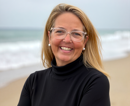 Smiling person in glasses and black turtleneck standing on a beach with ocean waves behind them