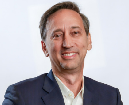 Studio headshot of a smiling person in a dark blazer and white shirt against a light background