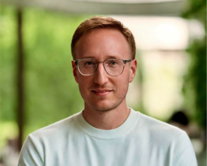 Portrait of a man with glasses in a light sweater, standing against a blurred green outdoor background