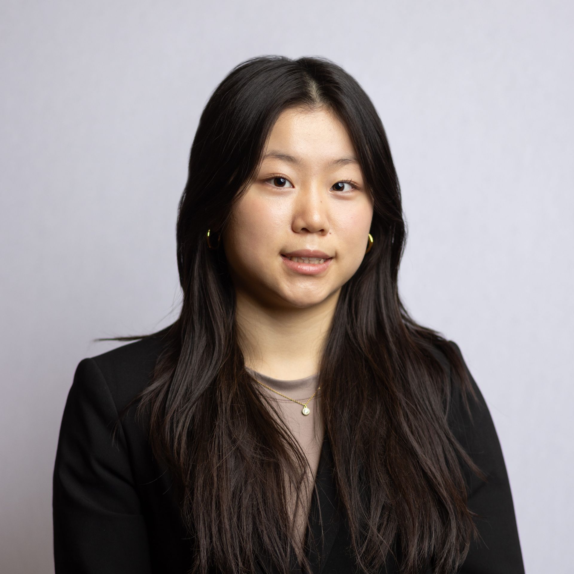 Studio portrait of a person with long dark hair wearing a black blazer against a light gray background