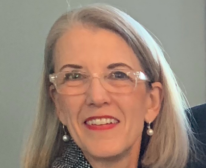 Smiling woman with gray hair, glasses, and pearl earrings against a neutral background
