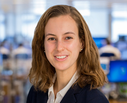 Smiling woman in a navy blazer in an office with blurred computer monitors in the background