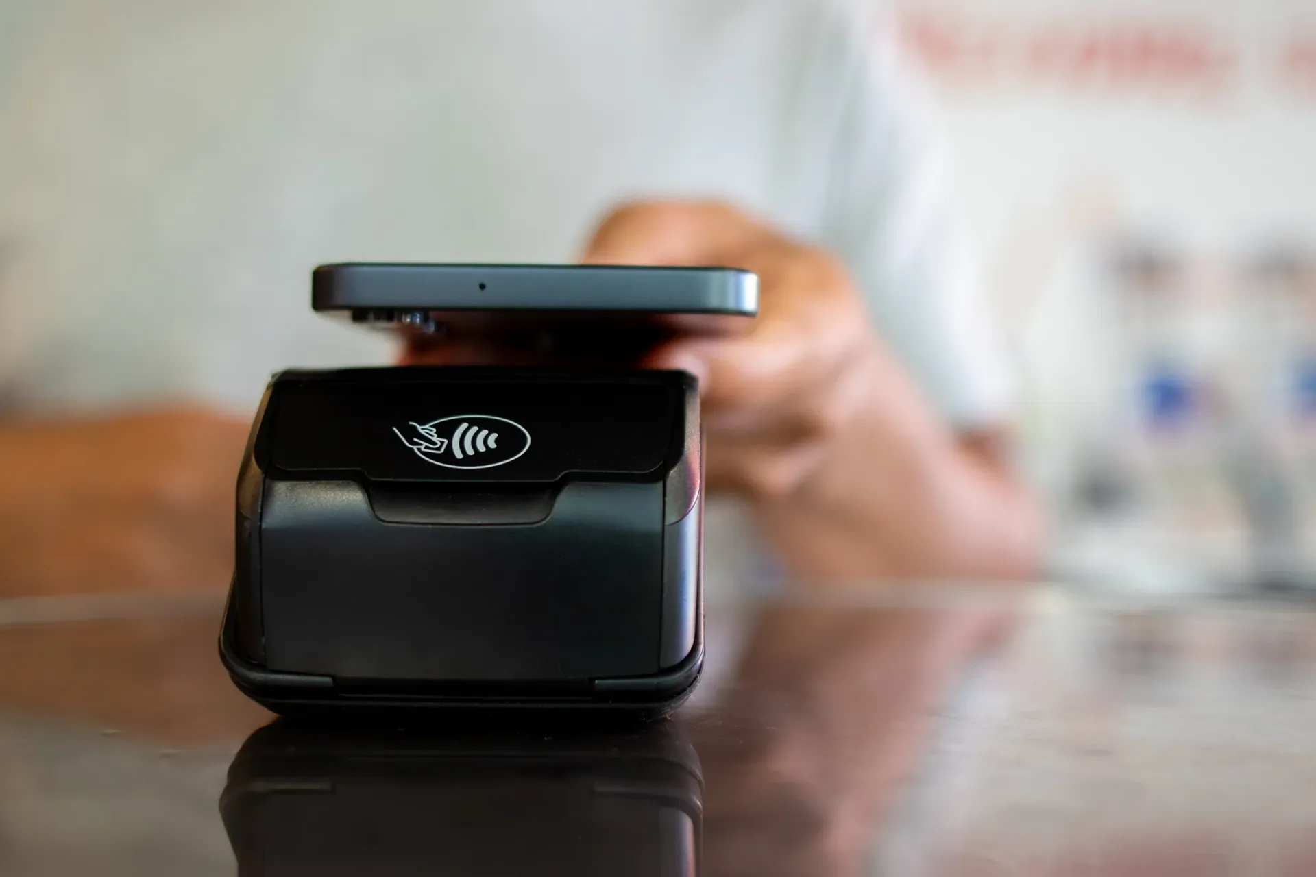 Black payment terminal on a counter with a person holding a phone in the blurred background