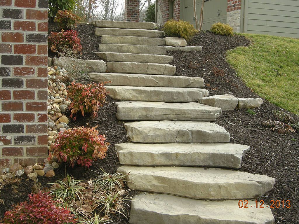 Stone steps leading up a sloped yard, surrounded by plants and mulch next to a brick wall.