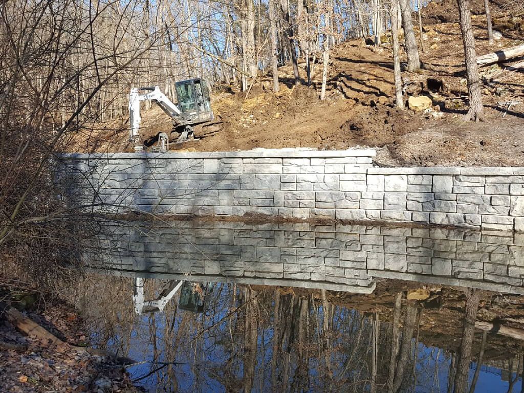Excavator working above a stone wall reflecting in a body of water. Trees line the banks.