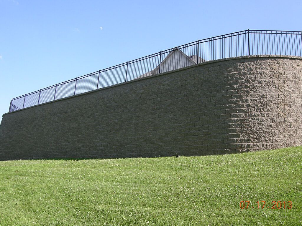 A retaining wall made of stacked blocks with a black fence on top, against a blue sky.
