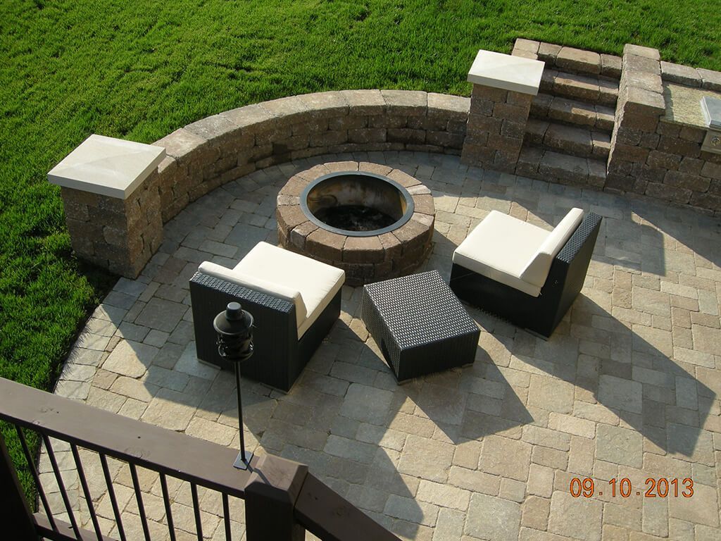 Patio with seating around a fire pit, surrounded by a low stone wall and stairs, on a sunny day.