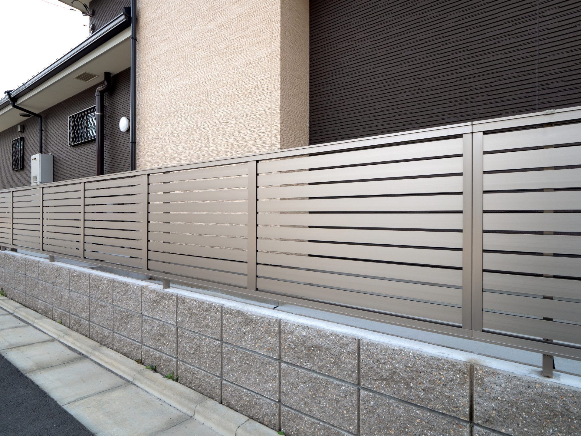 Tan metal horizontal slat fence atop a gray brick wall, adjacent to a sidewalk and building.