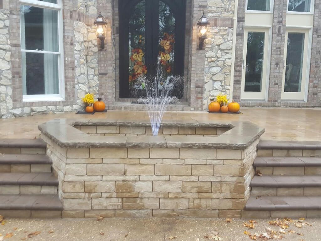 Stone fountain in front of a house, water spouting, flanked by pumpkins and stairs.