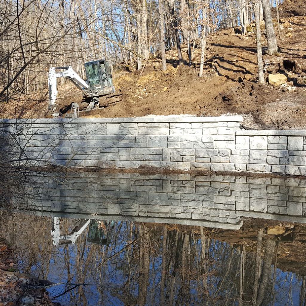 Excavator near a retaining wall and reflective water. Trees in the background; sunny day.