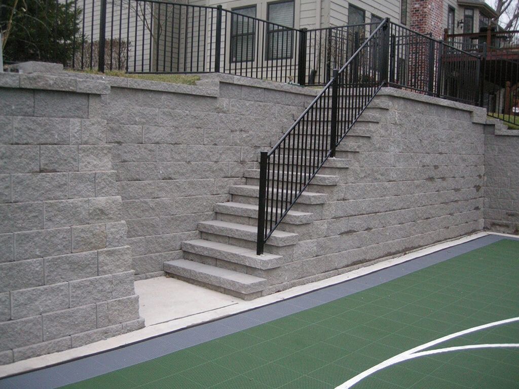 Stone retaining walls with stairs and a black handrail leading up to a black fence.