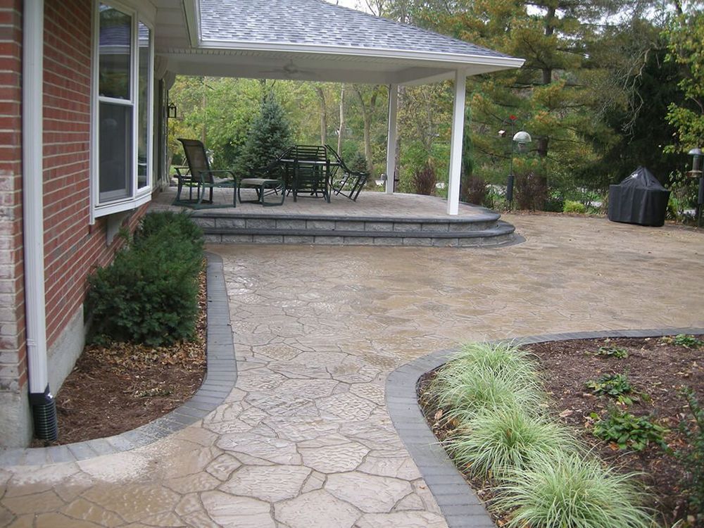 Stone patio with steps leading to a covered seating area near a brick house, bordered by plants and a landscaped yard.