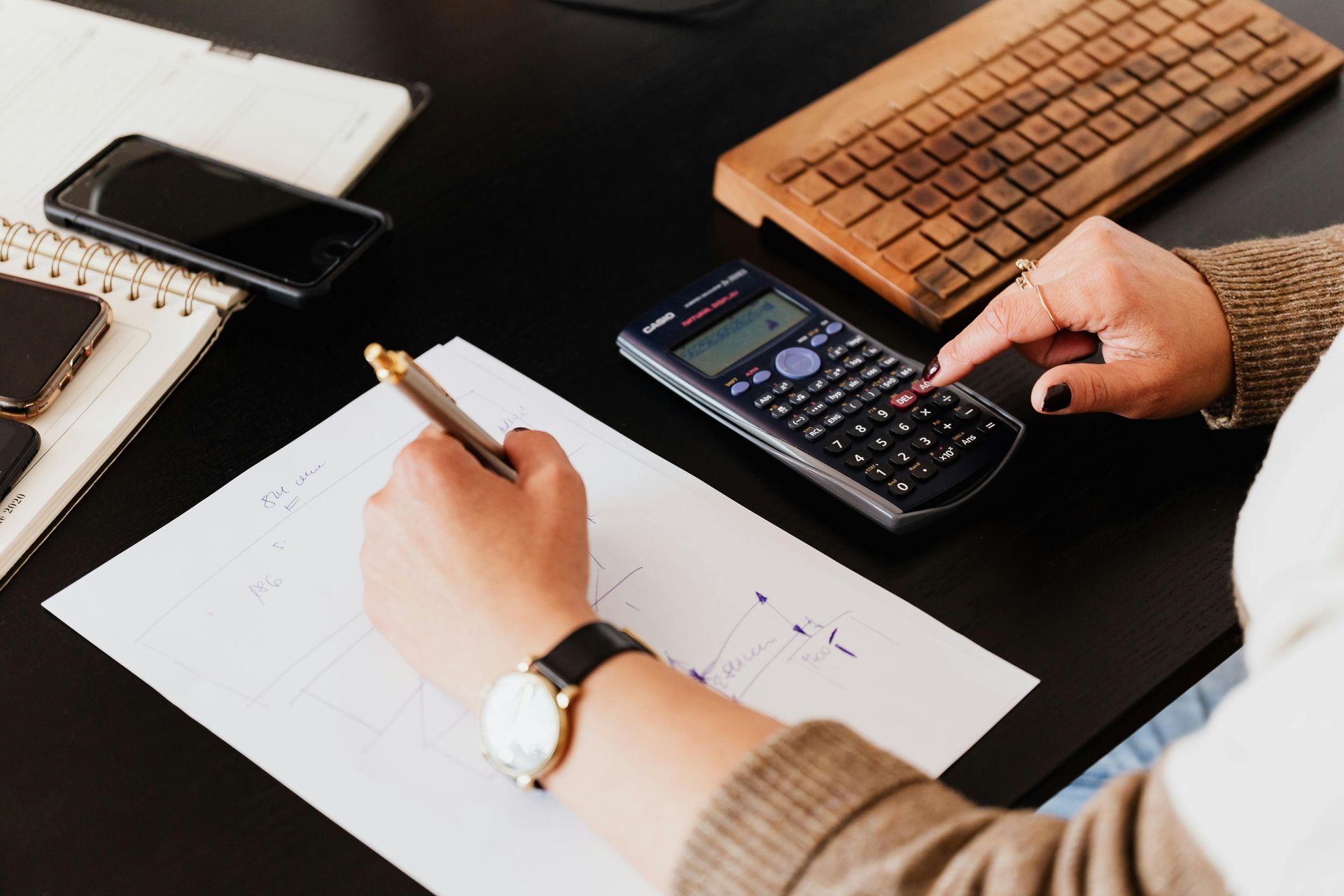 Person working on a desk, using a calculator and writing on paper, with a phone and keyboard.