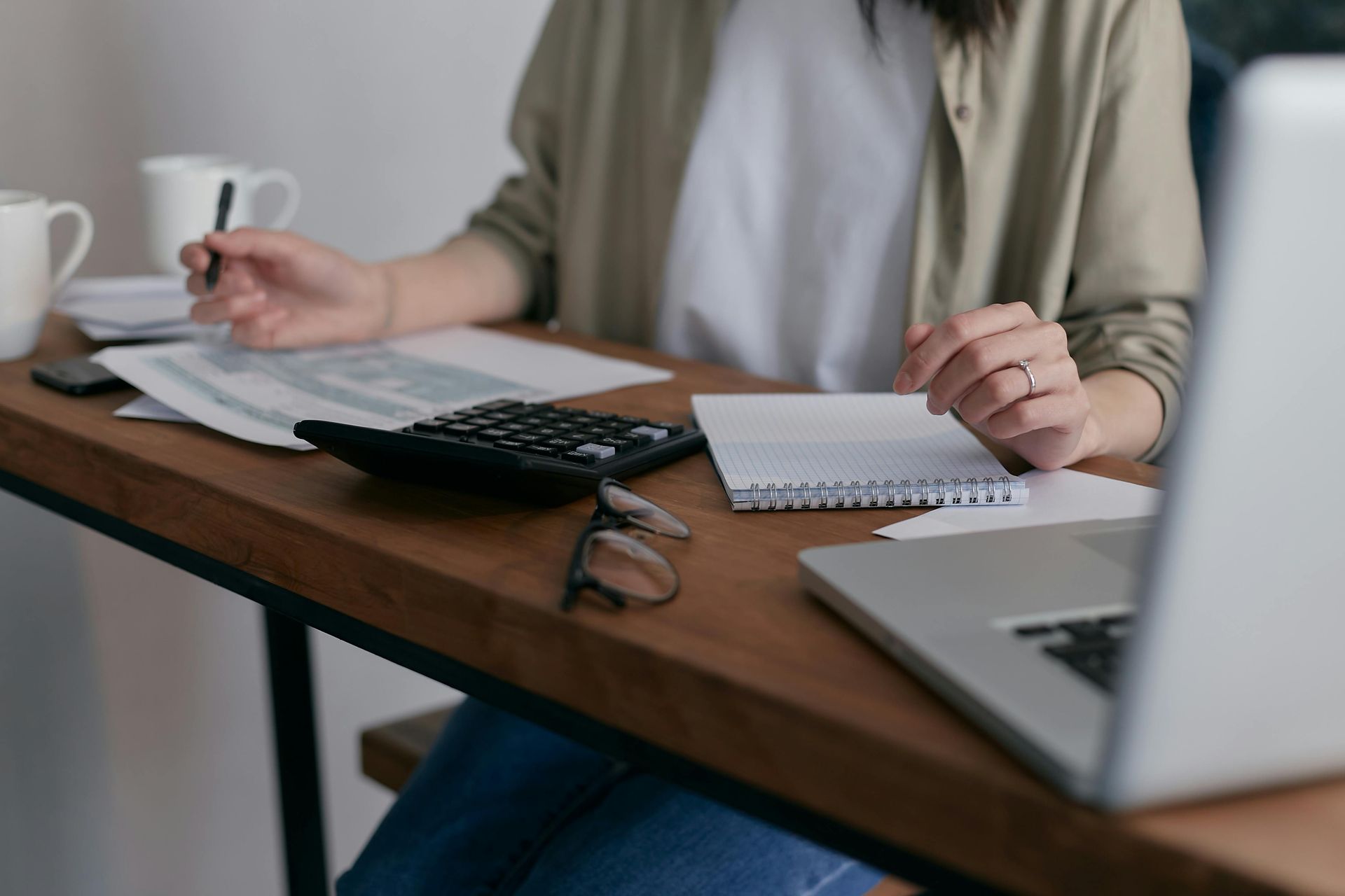 Woman sitting at desk doing finances with calculator, laptop, papers, and coffee.