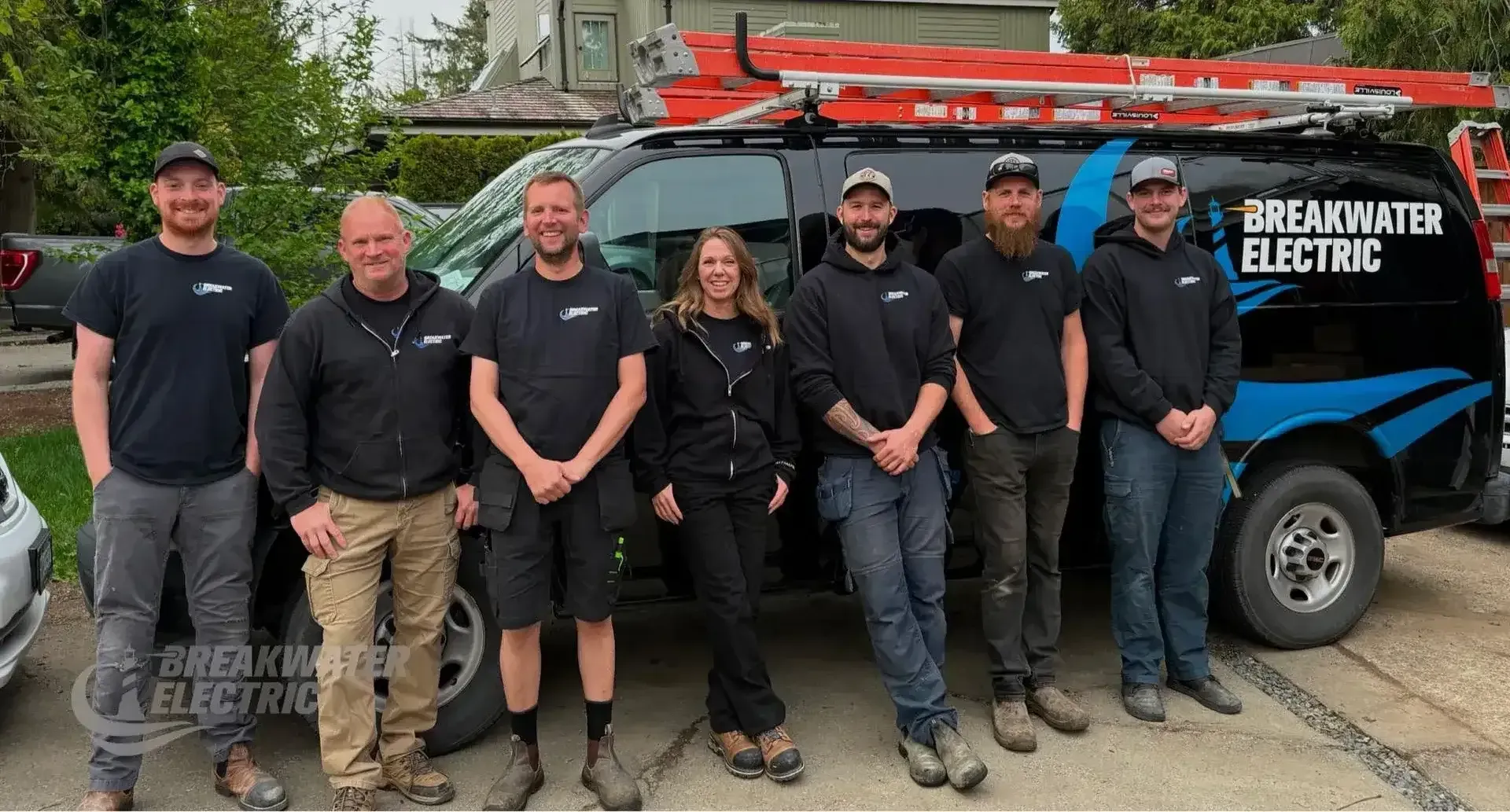 Six workers stand beside a white service van with ladders and tools in a driveway.