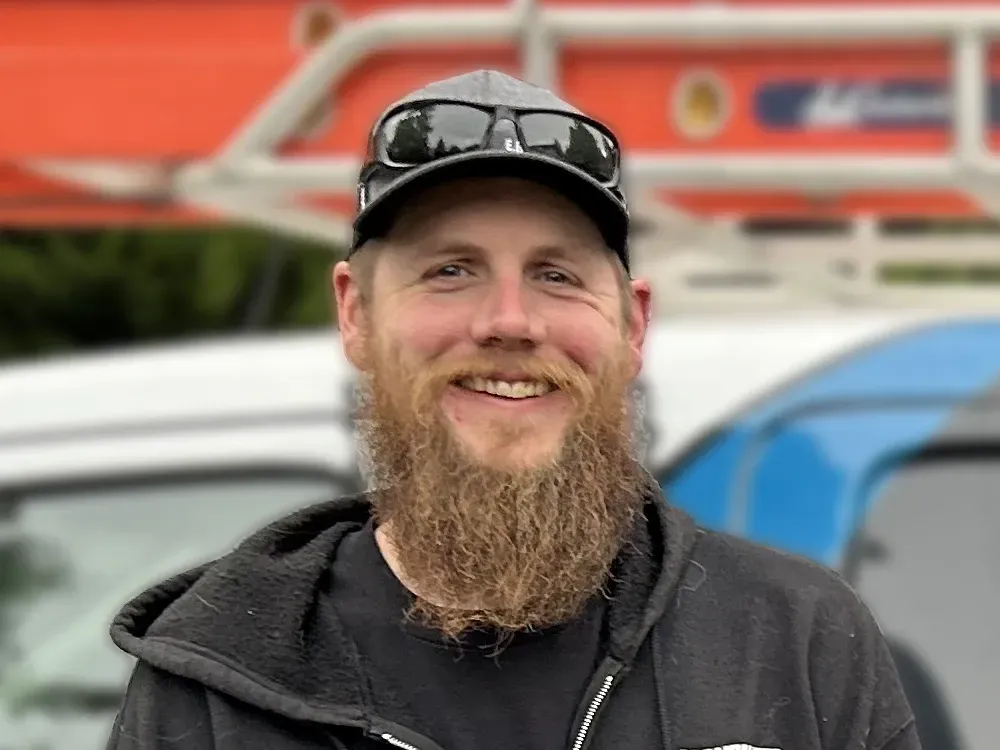 Smiling bearded man in a black cap and jacket standing outdoors by a vehicle and orange structure