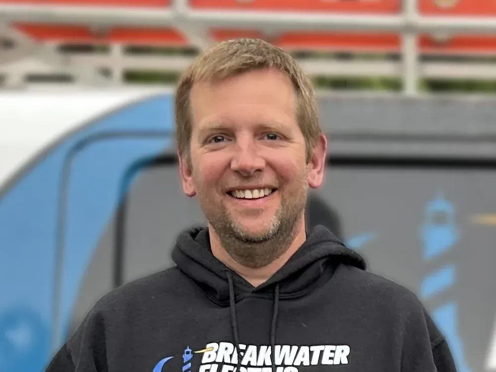 Smiling person in a black hoodie standing in front of a blue vehicle with blurred red and white background