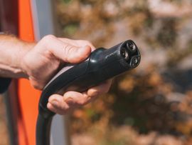 Hand holding a black EV charging connector near an orange vehicle body