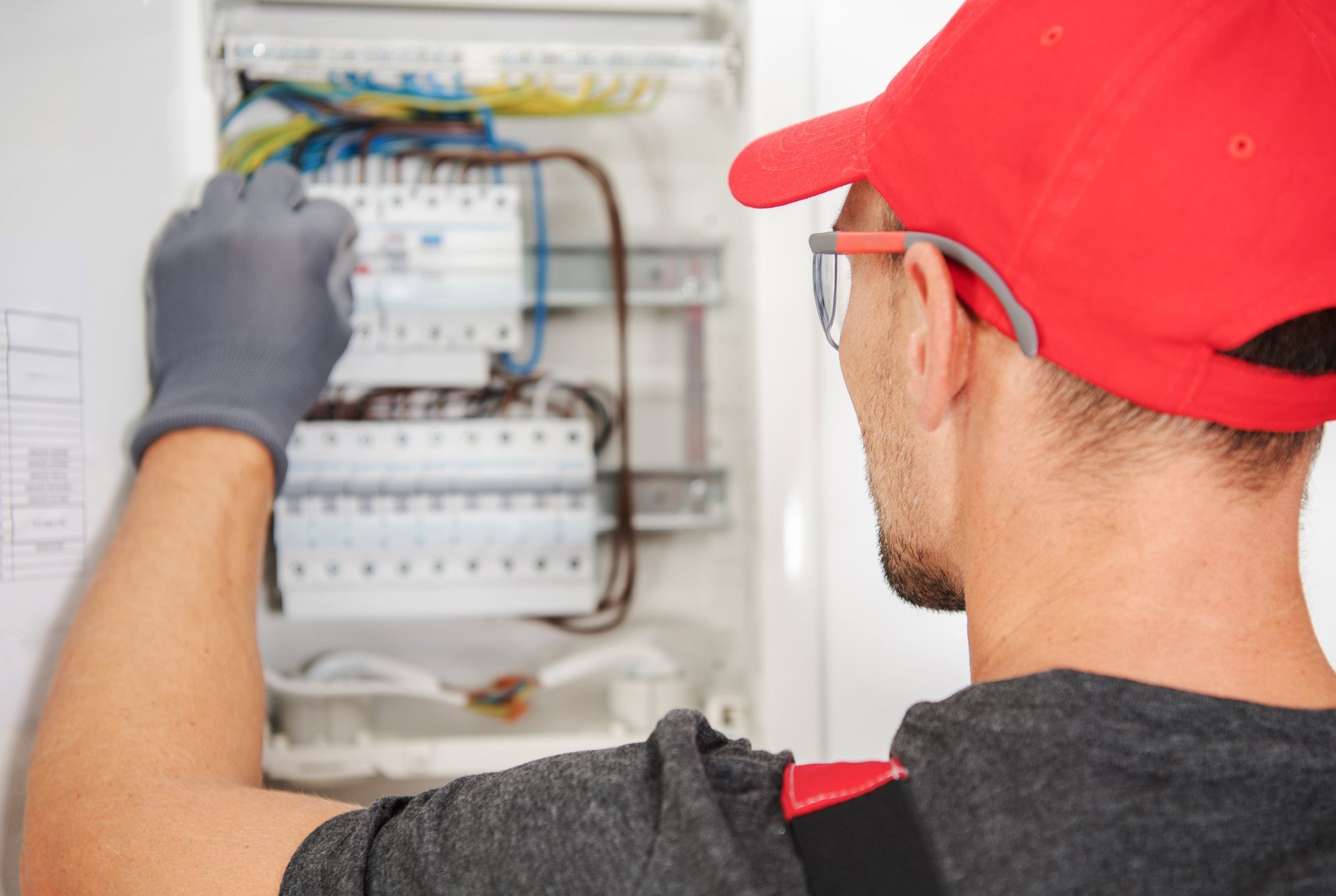 Electrician in a red cap and safety glasses working on a circuit breaker box.