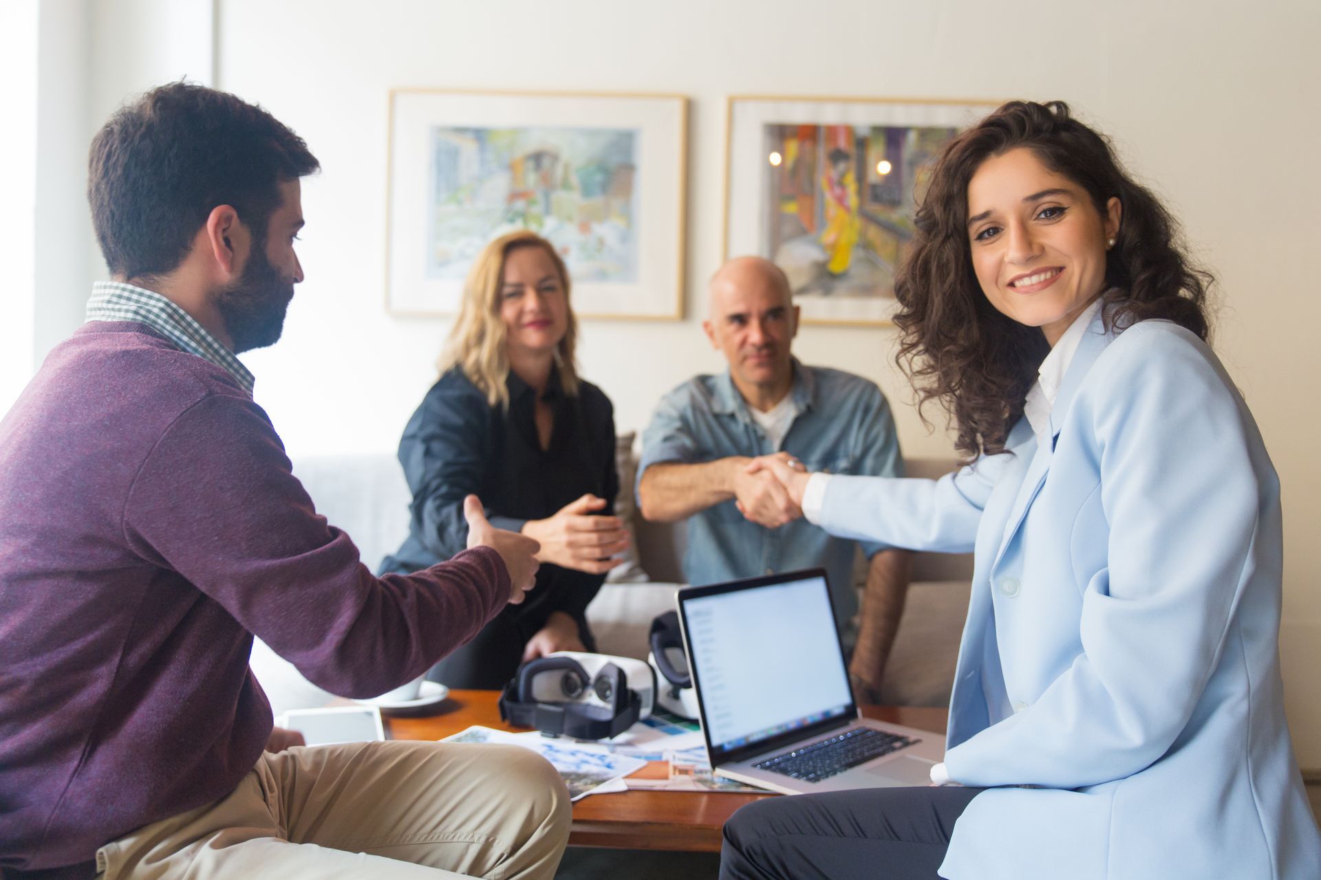 Business people shaking hands at a table, laptop, and papers present, others watch.