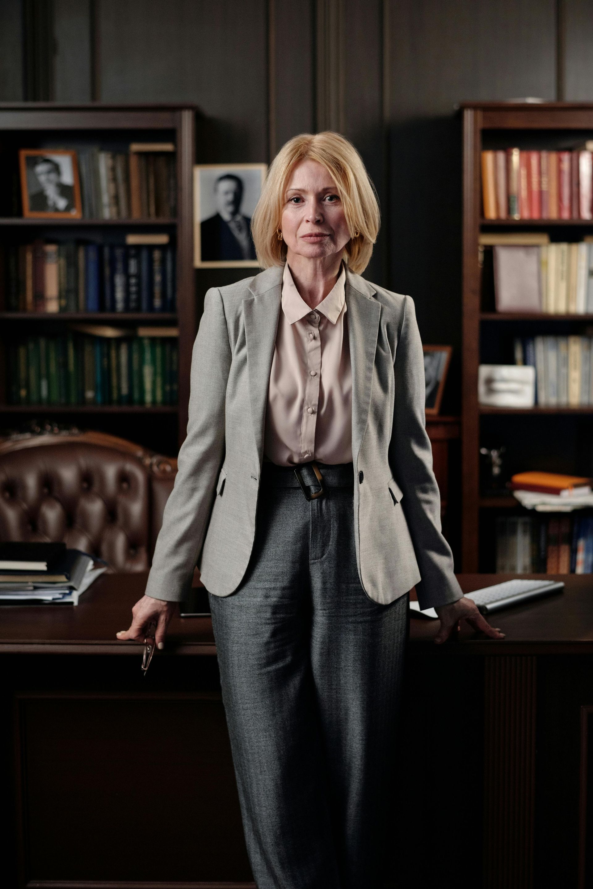 Woman in a gray blazer stands behind a desk in a wood-paneled office.