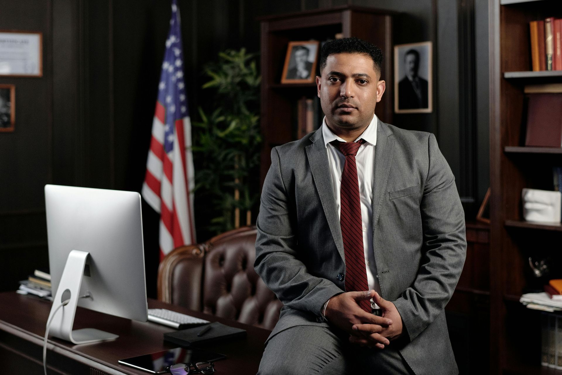 Man in a suit sits on a desk in an office with an American flag.