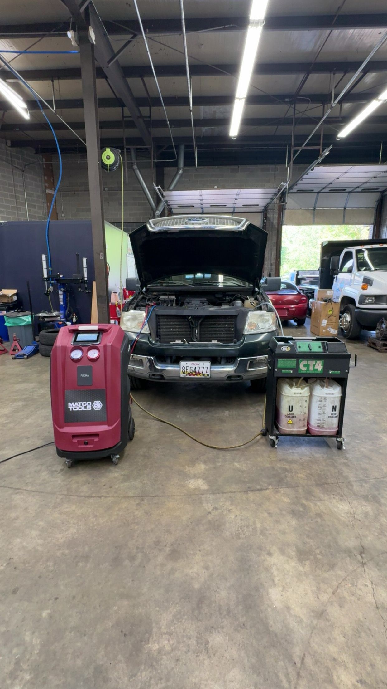 A red AC service machine and a fluid cart sit in front of a dark pickup truck with its hood open in an auto repair shop.
