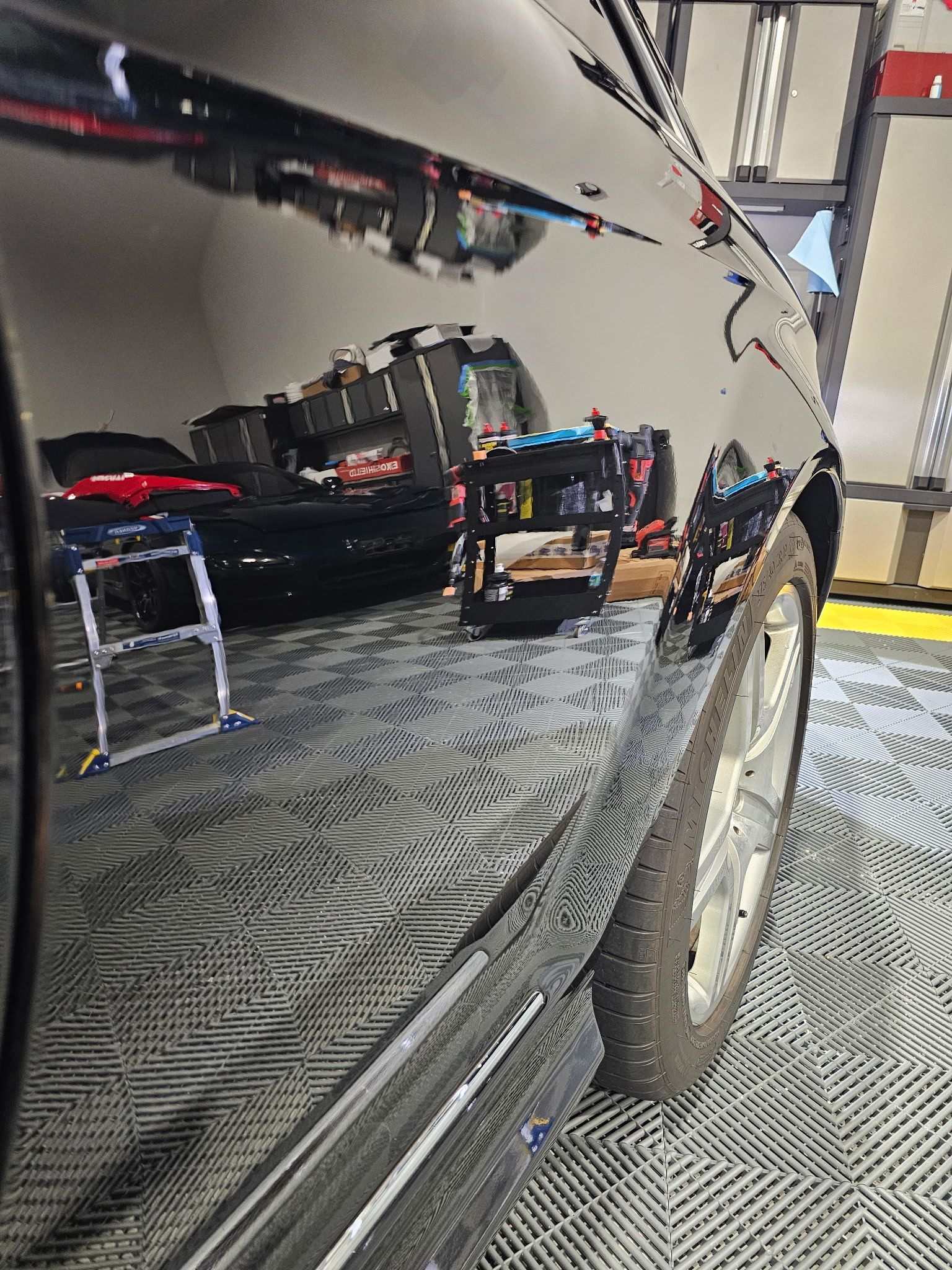 Black car side reflecting a workshop with tools and a shiny wheel.