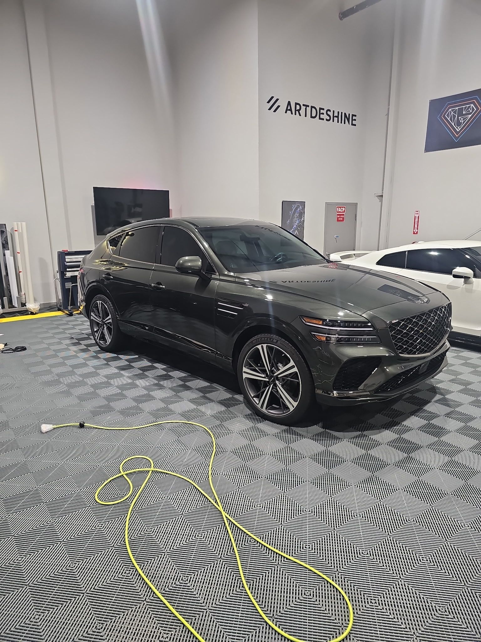 Dark gray Genesis GV70 SUV parked on a patterned garage floor in a showroom with equipment scattered nearby.