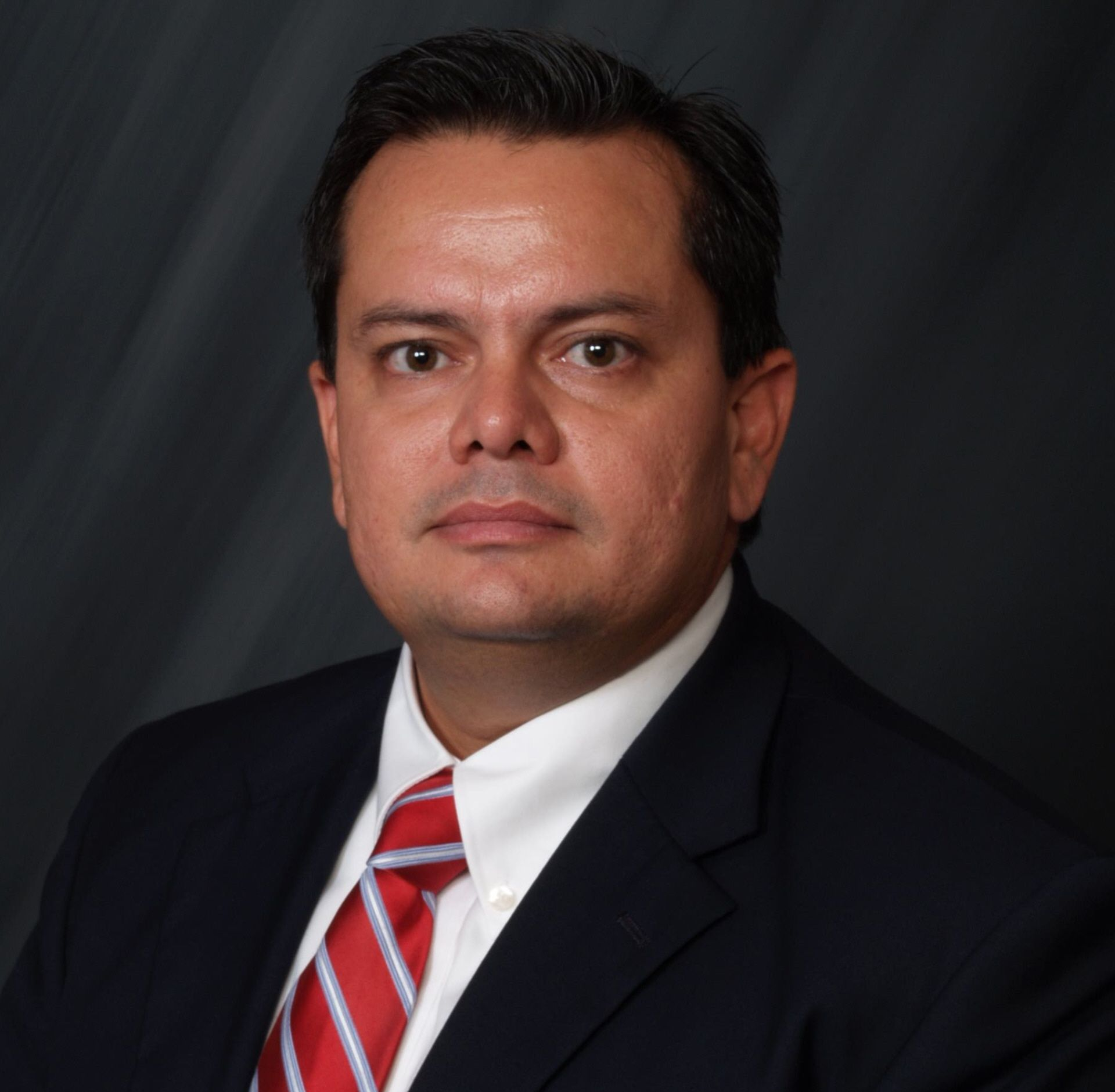 Studio headshot of a man in a dark suit, white shirt, and red striped tie against a dark background