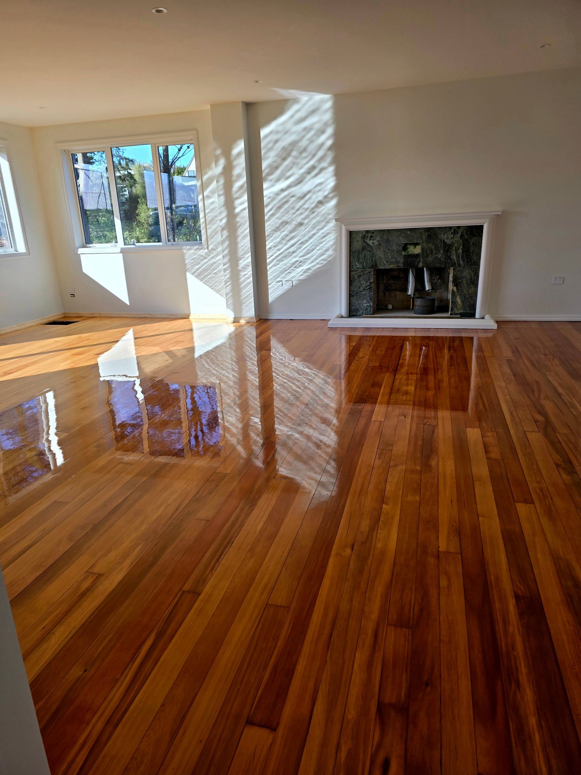 Empty room with glossy hardwood floors, white walls, windows, and a fireplace.