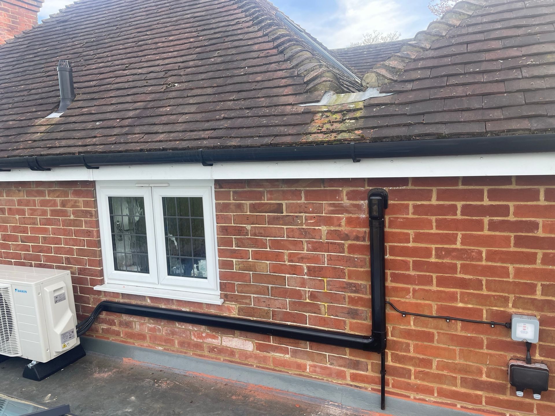 Brick house exterior with white window, rooftop moss, and black utility pipes along the wall
