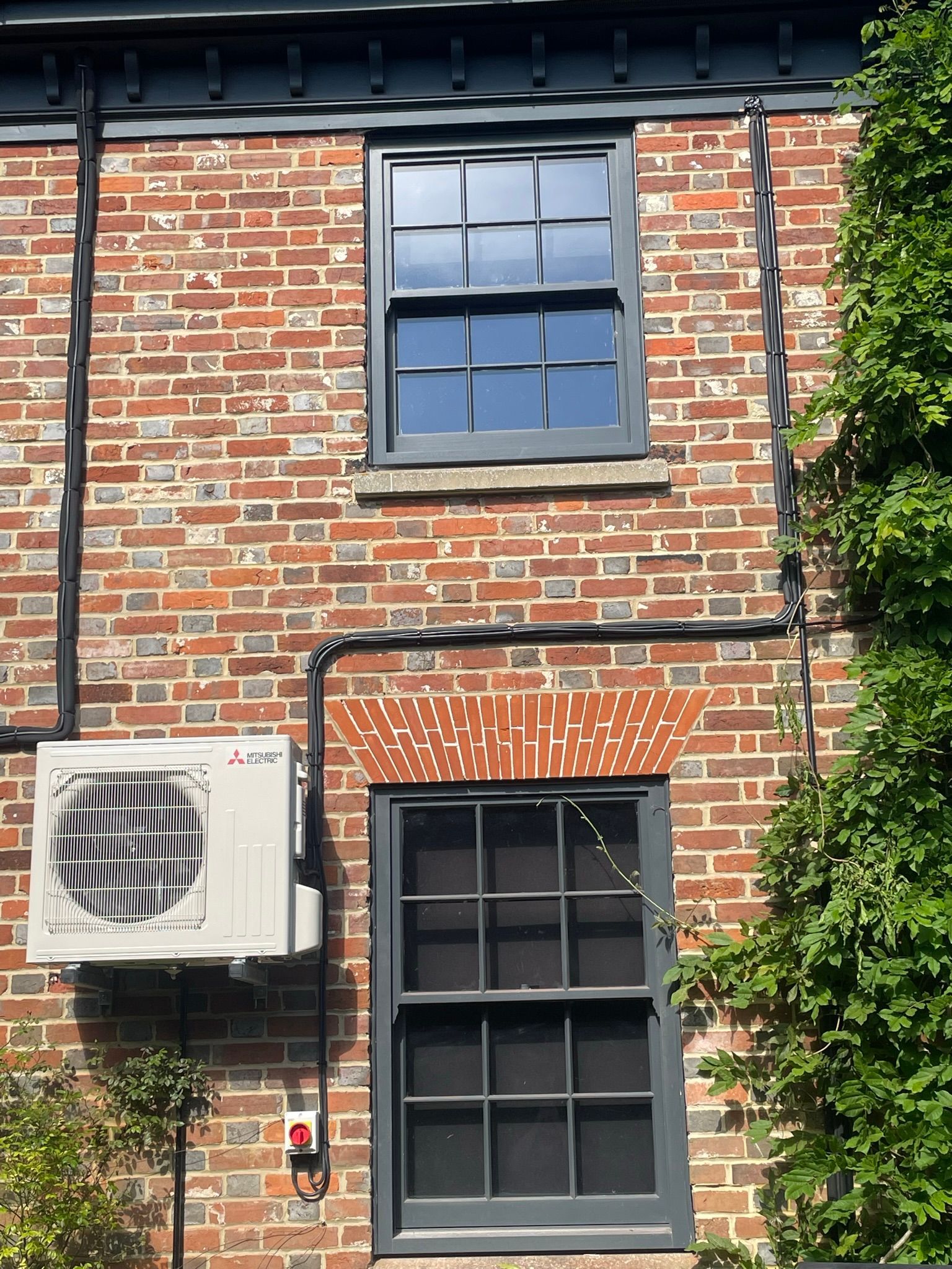 Red brick building facade with two windows, a wall-mounted air conditioner, and green ivy on the right.