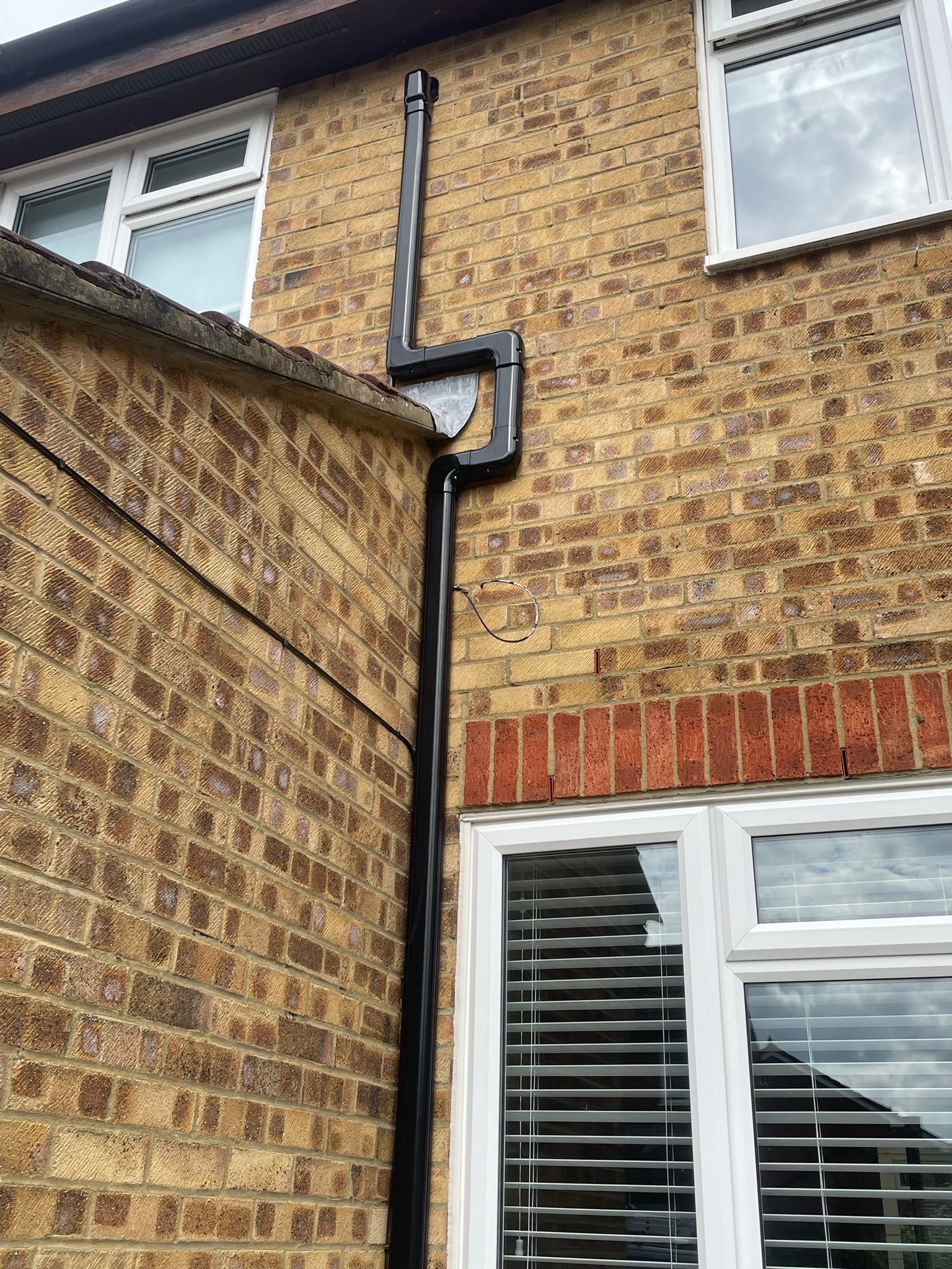 Brick house exterior with black downspout between a two-story wall and a lower roofline, white window below.