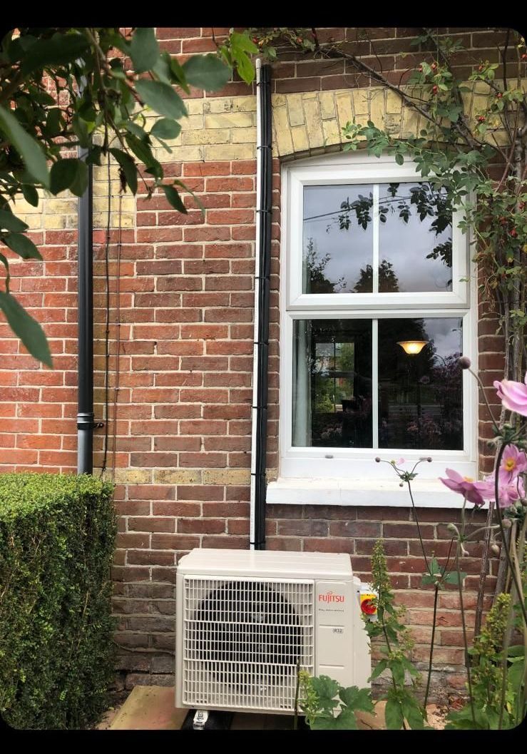 Brick house wall with white-framed window and an outdoor AC unit beneath green hedges.