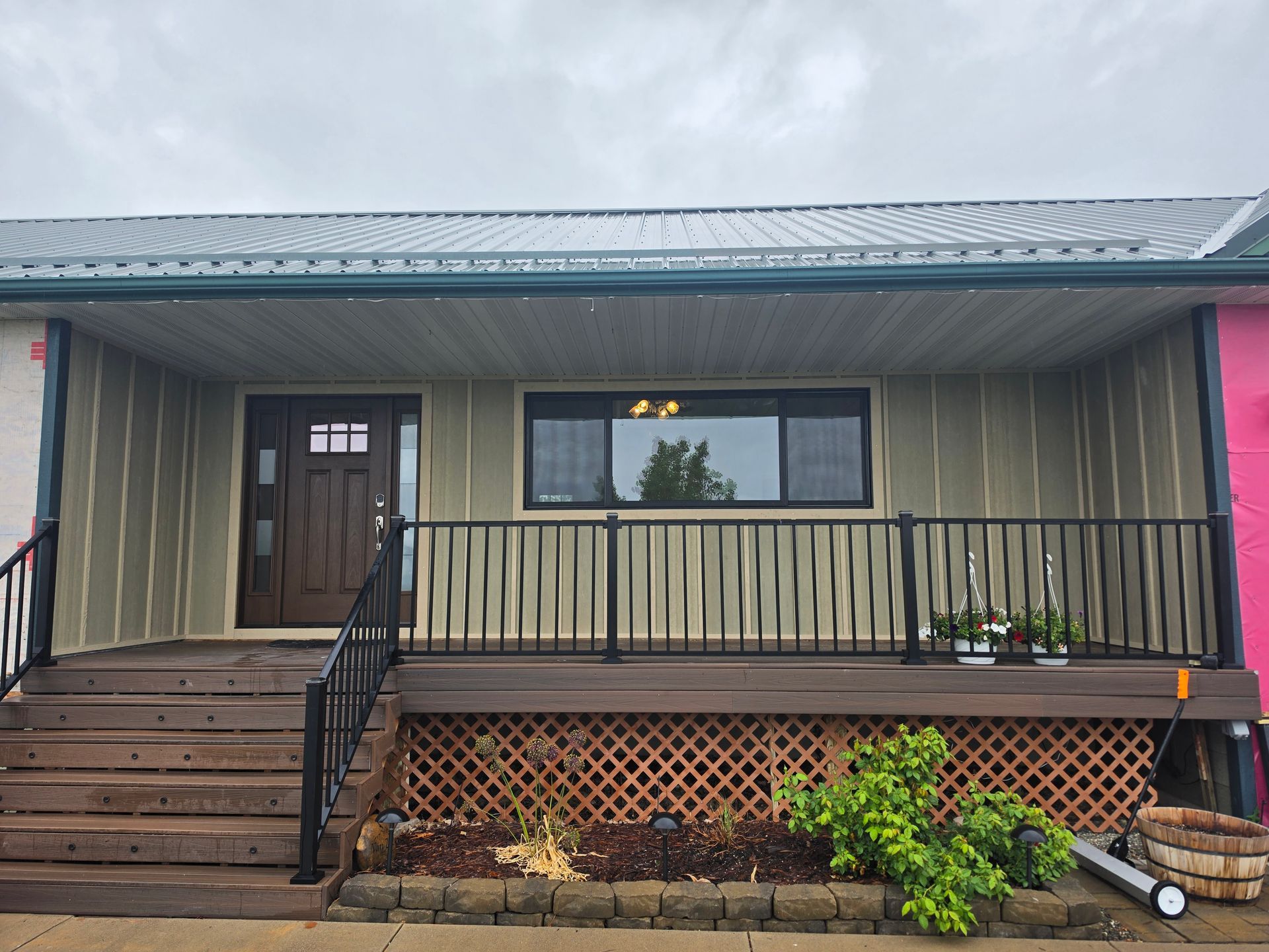 Front porch of a house with wooden steps, black railing, and beige siding under a metal roof.