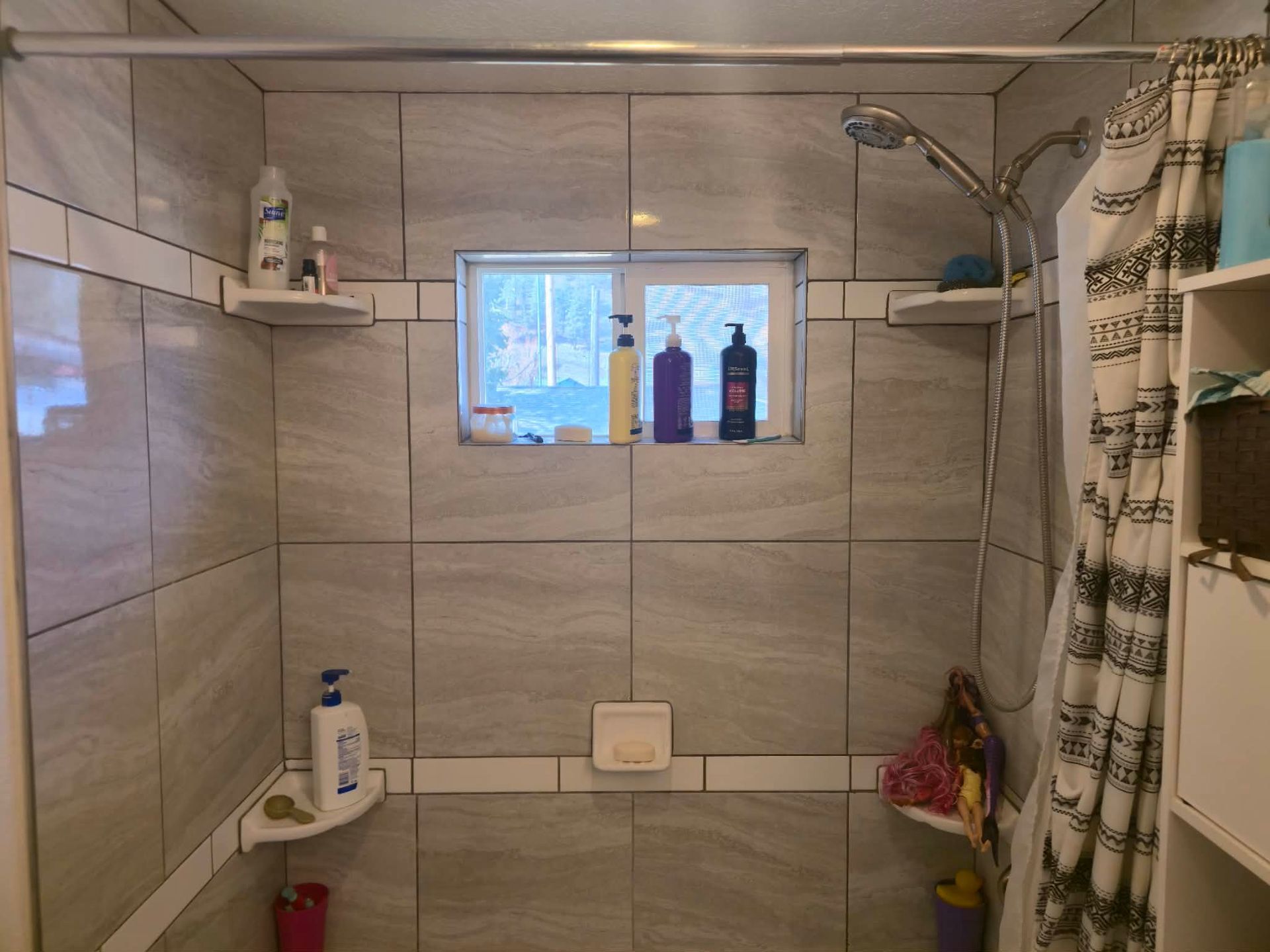 Bathroom shower with gray tile, window shelf holding bottles, and a plaid shower curtain on the right.