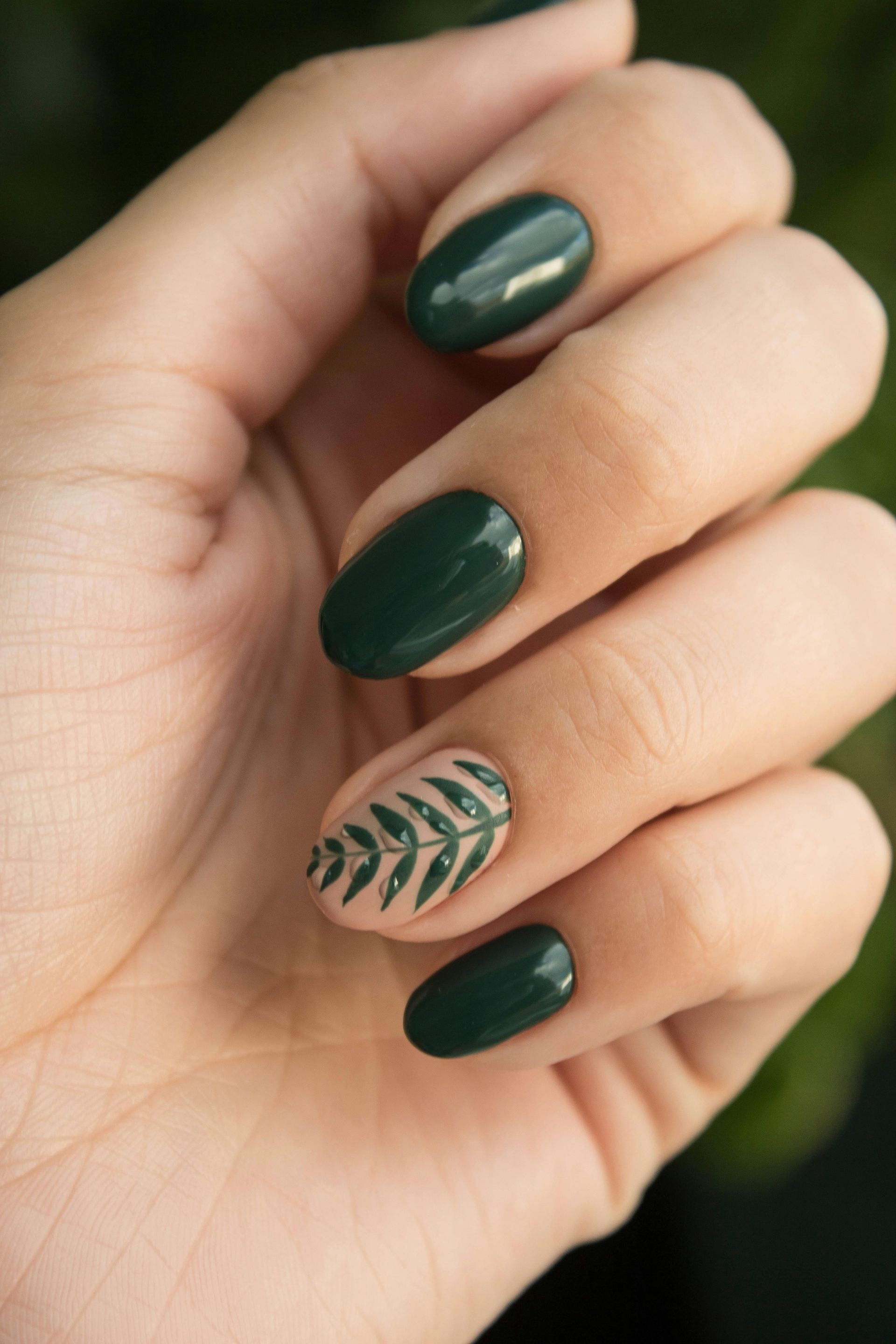 Hand with dark green polished nails; one nail features a beige fern design.
