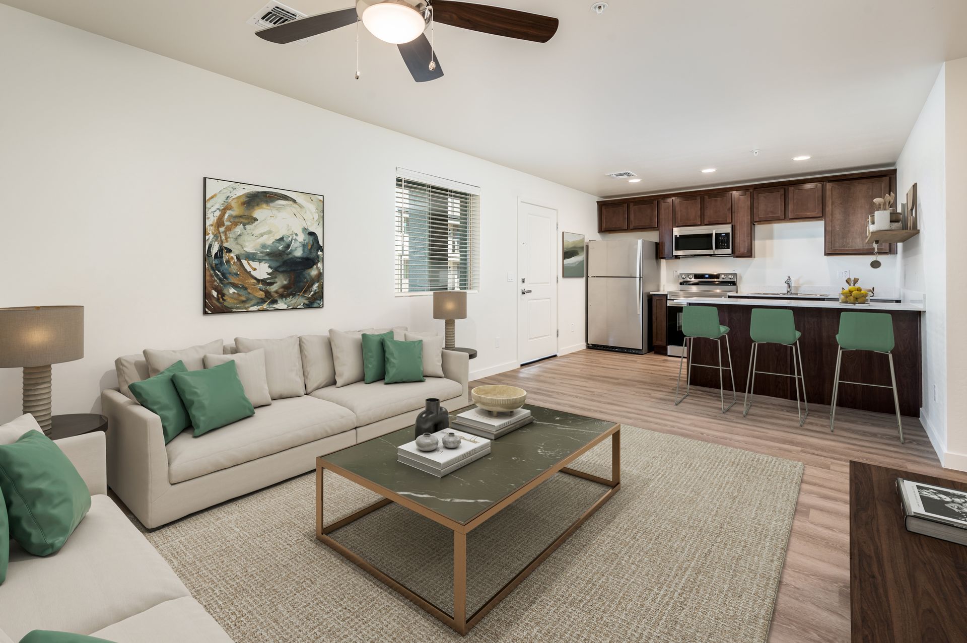 Cozy living room with kitchen; beige sofa, green pillows, brown cabinets, hardwood floors, and a ceiling fan.