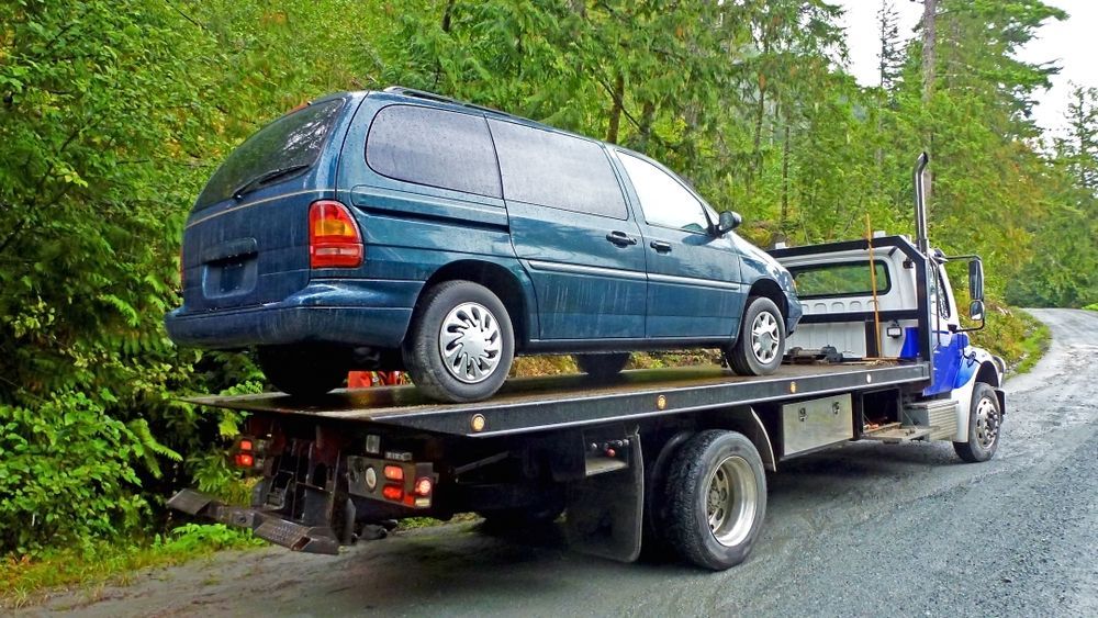 Dark Teal Minivan Transported On Flatbed Tow Truck Gravel Road Trees — Penfold Towing in Rainbow Beach, QLD