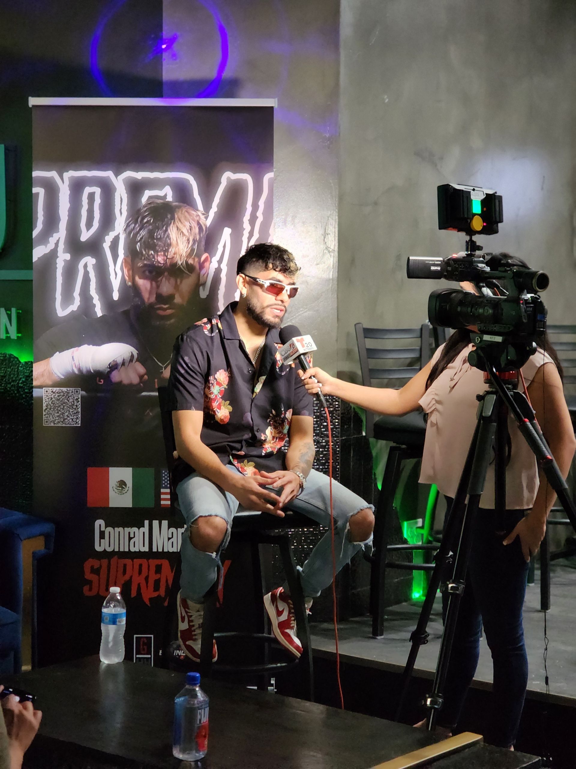 Man seated on a stool being interviewed on a small stage while a camera records him.