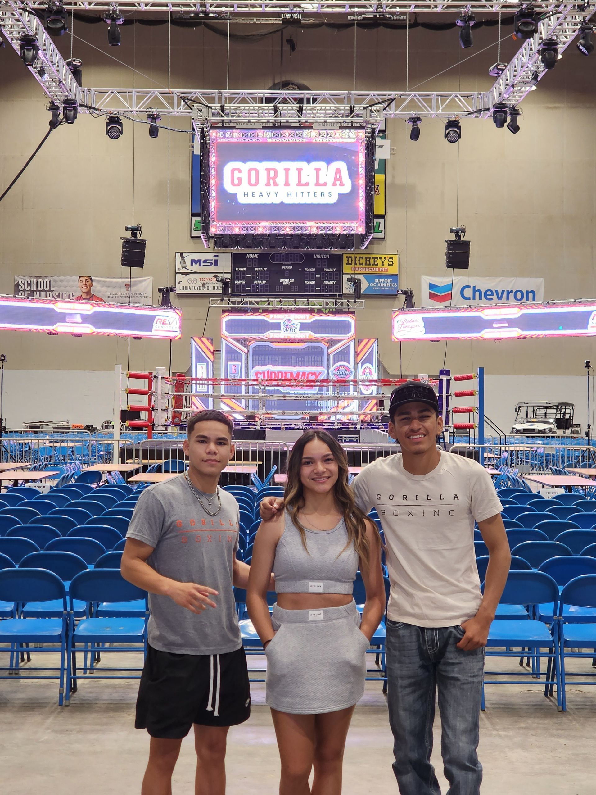 Three people pose in front of a large concert stage with blue chairs and a “Gorilla” screen.