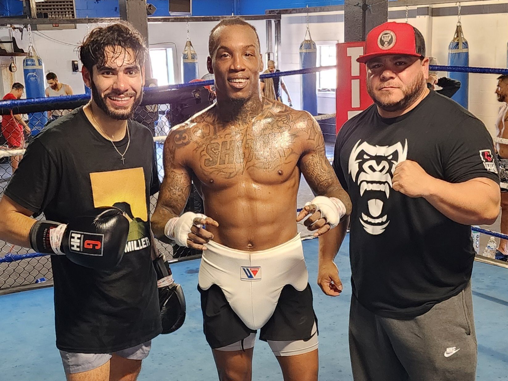 Three boxers pose in a gym, with the center fighter shirtless and glistening with sweat, flanked by two teammates.